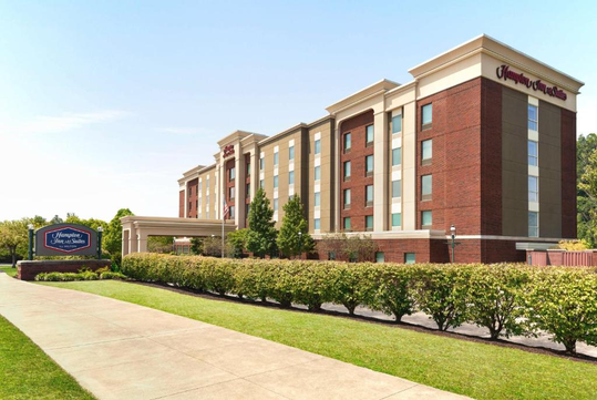 Four-story Hampton Inn & Suites hotel with red brick and tan exterior, landscaped bushes, and a large blue sign near the entrance.