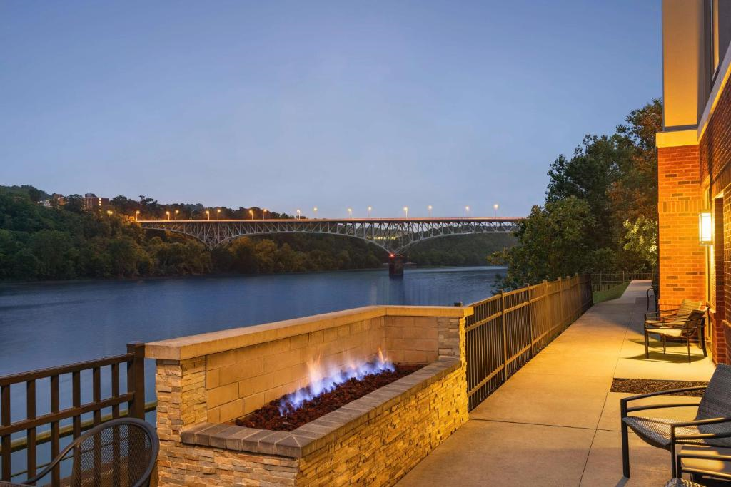 A riverside patio with a lit fire pit, chairs, and a view of a bridge spanning the water at dusk.