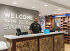 A hotel front desk staff member stands behind the reception counter, with a welcome sign and a snack shop visible in the background.