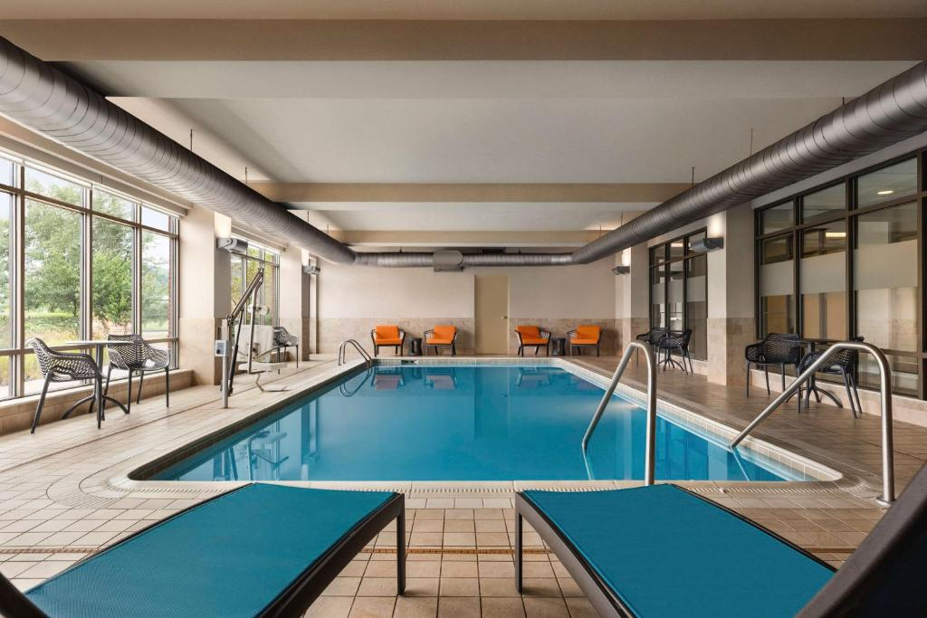 Indoor swimming pool with metal handrails, lounge chairs in the foreground, and orange chairs along the far wall. Large windows let in natural light.