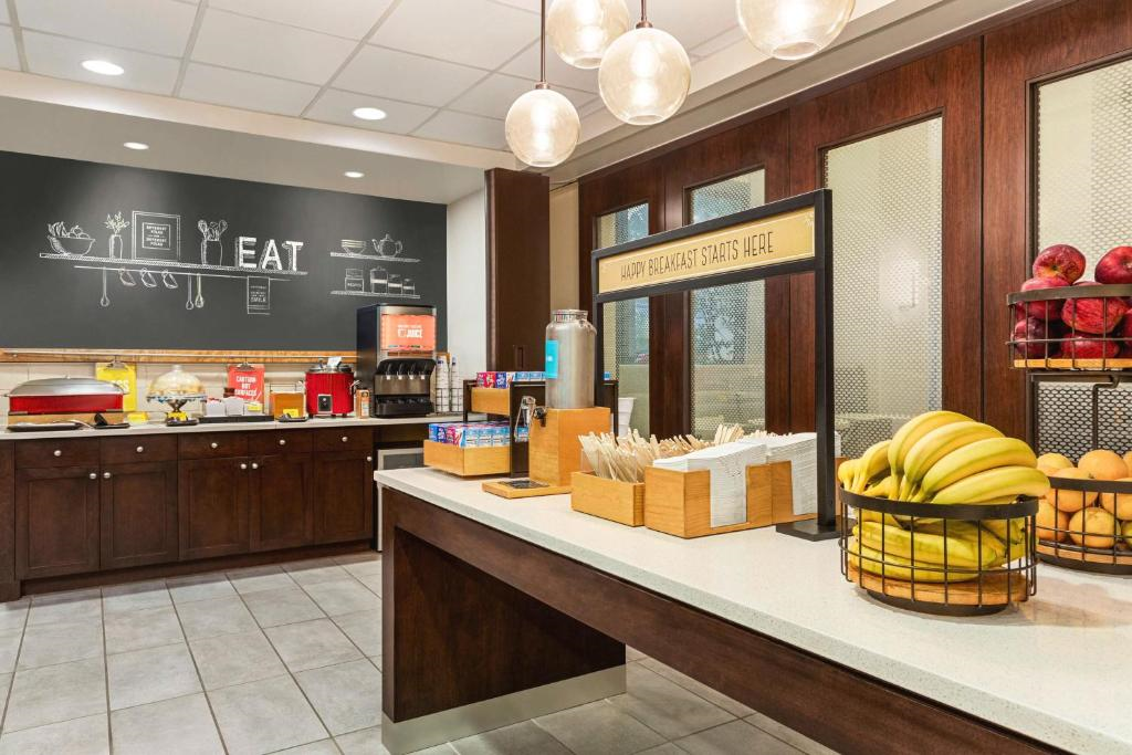 Hotel breakfast area with fruit, utensils, and drinks on the counter; food and beverage dispensers visible in the background with "EAT" sign on the wall.