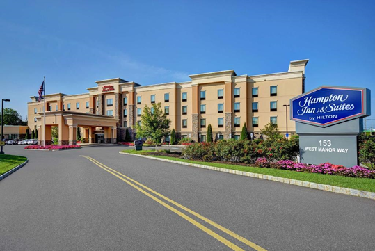 Exterior view of Hampton Inn & Suites with a landscaped entrance and prominent signage on the right. Road leads to the front of the building, with a flag visible near the entrance.