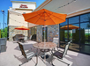 Outdoor patio with orange umbrellas at a Hampton Inn & Suites. There are tables and chairs on the concrete surface, adjacent to the hotel's glass doors and stone walls.