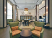 Spacious hotel lobby with high ceilings, modern chandeliers, armchairs, a central table, and seating areas. Neutral color palette with accents of green and orange patterns.