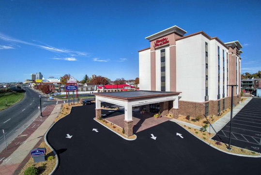 Large hotel building with a red and white exterior, entrance canopy, and nearby parking lot. Surrounding buildings and a road are visible under a clear blue sky.