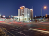 Hotel building illuminated at night with cars passing by, leaving light trails. Parking lot in front and surrounding streetlights visible.