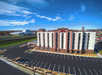 Aerial view of a multi-story hotel with a large empty parking lot. The building has brick and beige paneling. A white dome structure is visible in the background under a clear blue sky.