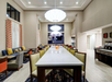 Modern communal area with a long table, chairs, and sofas. Hanging lights illuminate the space. A TV and coffee station are in the background. Neutral color palette with red accents.