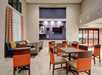 Hotel lobby with tables and chairs, a blue sofa, and mounted TV. Curtains with red patterns cover tall windows. The space is bright with neutral tones.