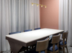A meeting room with a long white table, six chairs, a pink wall, sheer curtains, and a modern overhead light fixture.