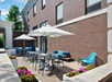 Outdoor patio with tables, chairs, and umbrellas in front of a brick building. Colorful flowers in planters line the walkway.