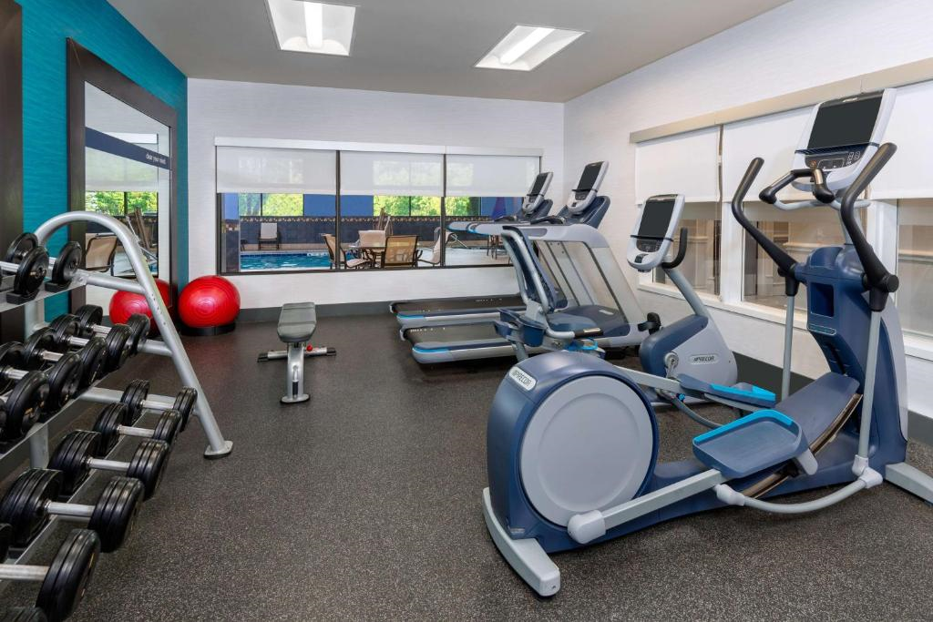 A gym with treadmills, an elliptical, stationary bikes, free weights, two red exercise balls, and a bench on a rubber floor. Large windows provide natural light.