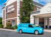 A blue Hampton Inn van is parked in front of the hotel's brick and white facade on a clear day.
