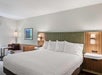 Hotel room with a large bed, four pillows, two bedside tables with lamps, a sofa, a side table, and a wall picture. Neutral color scheme with wood accents.