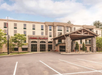 Four-story hotel exterior with a covered entrance and empty parking lot, set against a cloudy sky.