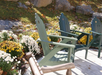 Three Adirondack chairs on a sunny patio surrounded by white and yellow flowers, with rocks and grass in the background.