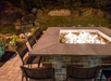 Outdoor patio with a square stone fire pit surrounded by wicker chairs, set against a backdrop of rocks, plants, and illuminated flowers.