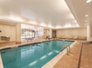 Indoor swimming pool with clear water, surrounded by lounge chairs and a table, in a well-lit room with beige walls and a white ceiling.