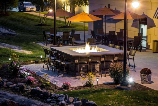 Outdoor patio with a square fire pit surrounded by wicker chairs, tables with orange umbrellas, and landscaping with flowers and rocks. A car is parked nearby, and a building wall is visible.