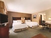 Hotel room with two double beds, white bedding, a flat-screen TV, a desk, and a lamp. There's a doorway leading to a bathroom area. The walls are beige and the carpet is brown.