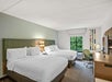 Hotel room with two double beds, a desk with a chair, a wall-mounted TV, and a window with a view of greenery. Modern decor with neutral tones.
