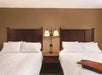 Two neatly made double beds with white linens are in a hotel room. A wooden nightstand with a dual-light lamp is positioned between the beds.
