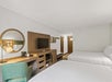 Hotel room with two double beds, a flat-screen TV on a wooden desk, a round mirror, a green chair, and framed art on the wall.
