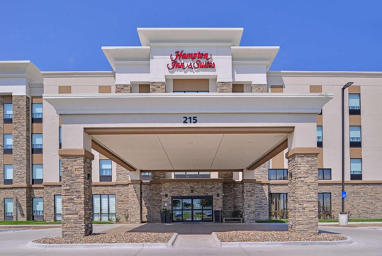 Front view of a Hampton Inn & Suites hotel entrance, featuring a covered driveway with stone pillars, beige exterior, and the number 215 displayed above the entrance.