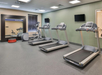 A gym room with three treadmills, an elliptical machine, a set of red exercise discs, a large mirror, and two wall-mounted TVs. The floor is carpeted, and there is a window with closed blinds.