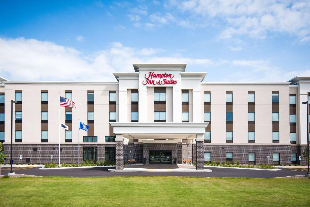 Front view of a Hampton Inn & Suites hotel with a clear sky above, featuring a modern facade, flags, and a circular driveway entrance.