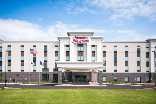 Front view of a Hampton Inn & Suites hotel with a clear sky above, featuring a modern facade, flags, and a circular driveway entrance.