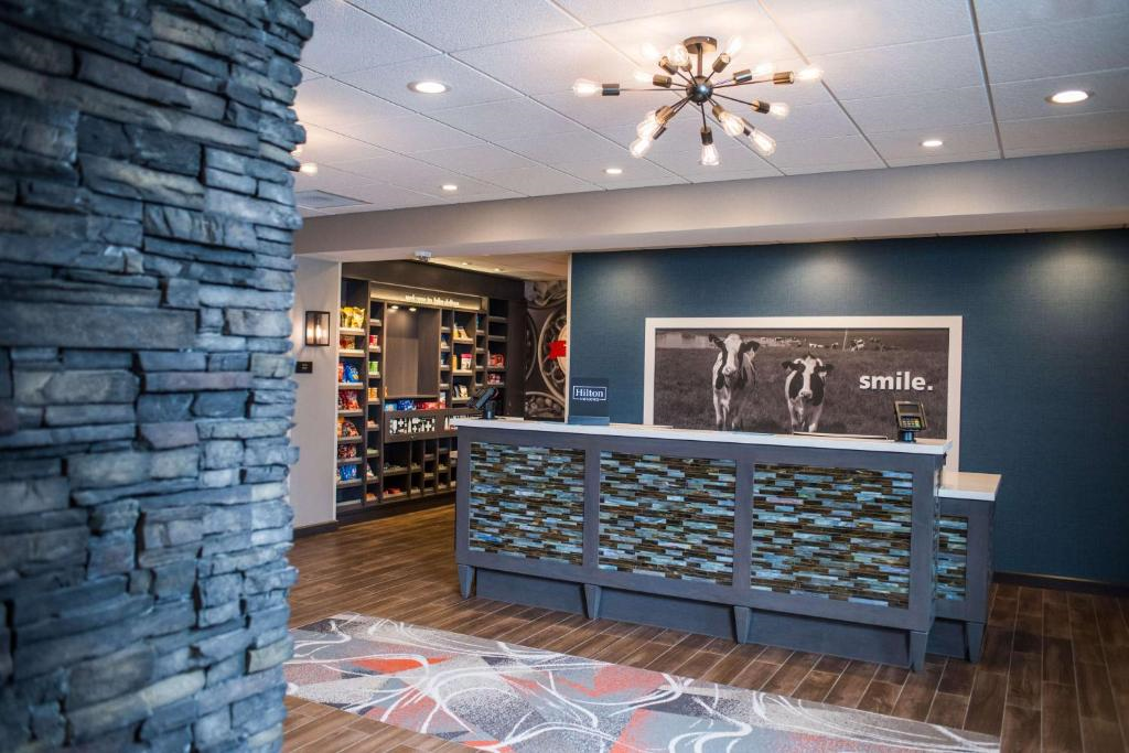 Hotel lobby with a modern design, featuring a stone column, front desk with decorative panels, wall art of cows with the word "smile," and a light fixture above. Shelves with items are in the background.