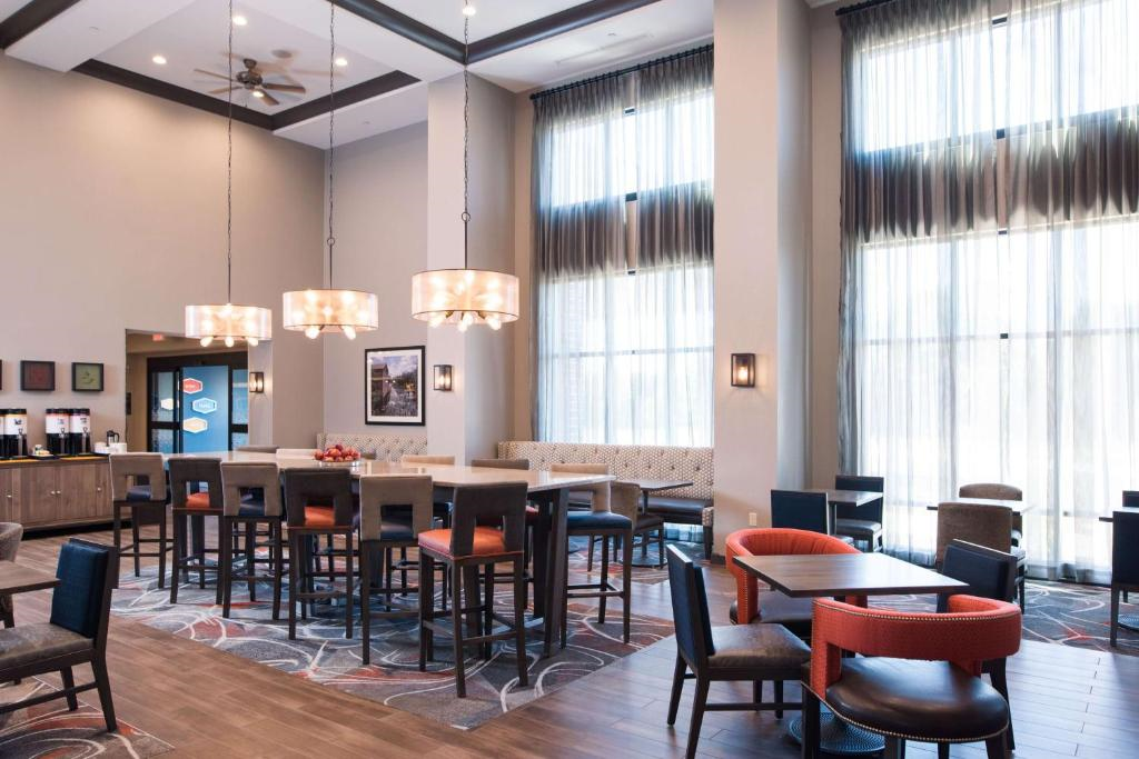 A modern hotel dining area with tall windows, multiple tables, high chairs, and hanging pendant lights. Walls are decorated with framed art.