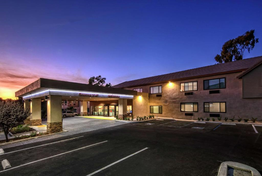 Modern hotel exterior with illuminated entrance at dusk. Two-story building, empty parking lot, and clear sky transitioning from sunset to night.