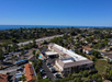 Aerial view of a coastal town with houses, trees, and roads, including a highway. The ocean is visible in the background under a clear blue sky.