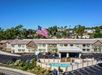 Aerial view of a hotel with a large American flag, parking lot, and outdoor pool, surrounded by trees and houses in a hilly area.