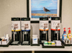 Four beverage dispensers labeled Light Roast, Dark Roast, Decaf, and Hot Water with cups, creamers, sugar packets, and stirrers in front. A framed picture of a seagull is on the wall.