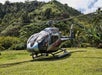 A silver and black Maverick helicopter parked in some grass in the Hana Rainforest with a mountain in the background on a sunny day.