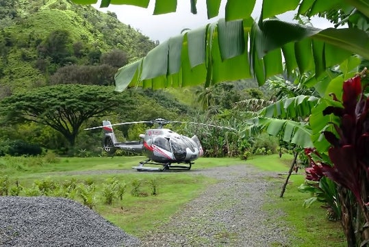 A silver and black Maverick helicopter parked in a gravel patch with a mountain behind them in the Hana Rainforest.