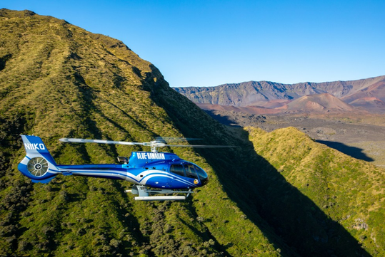 A blue helicopter flies over green, rugged hills with a mountainous landscape and clear blue sky in the background.