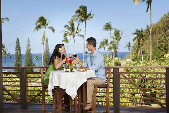 A couple sits at a table on a balcony, enjoying drinks and a meal with a view of palm trees and the ocean in the background.