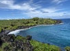 A curved coastline with black volcanic rocks, green vegetation, and clear blue water under a partly cloudy sky.