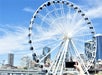 A large Ferris wheel in front of city buildings on a sunny day; a hand holds up a smartphone to take a photo of the scene.