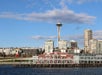 A waterfront building with a "70" sign appears in the foreground, with the Seattle skyline and Space Needle in the background under a blue sky with scattered clouds.
