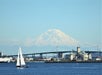 A sailboat on the water with an industrial bridge and buildings in the background, and a snow-capped mountain rising in the distance under a clear sky.