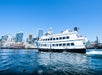 A white passenger ferry sails on the water with a city skyline, including a Ferris wheel, visible in the background under a clear blue sky.