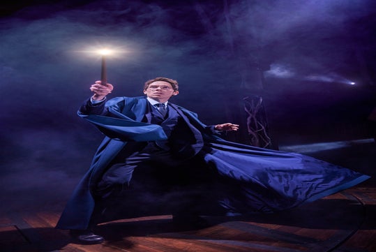 Person in a blue robe dramatically holds a lit wand amid swirling fog on a dimly lit stage.