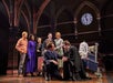 A group of people in a theatrical stage setting, wearing costumes with a mix of casual and formal attire, gather around a large trunk under a clock in an arched hall.
