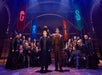 A group of people in robes gather on stage. A man holds a hat over another person's head, with colorful banners in the background.