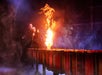 Two actors on stage near a flaming cauldron, surrounded by smoke and dramatic lighting.